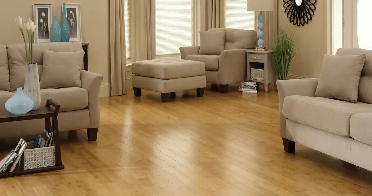 A living room with professionally installed bamboo flooring, beige sofas, a matching ottoman, a side table with a blue lamp, vases, framed pictures, and large windows with blinds and curtains in Wichita, KS