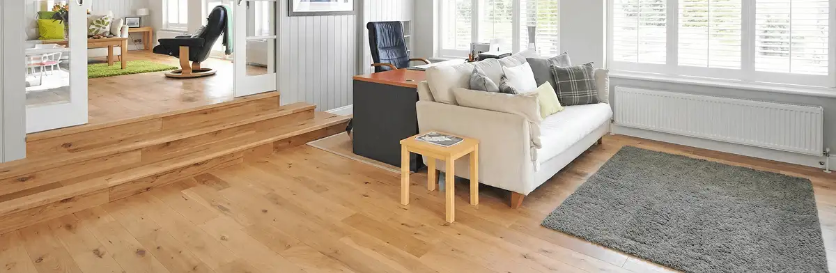A modern living room with professionally installed laminate flooring, a beige sofa, a small side table, a gray rug, and large windows with white shutters in Wichita, KS